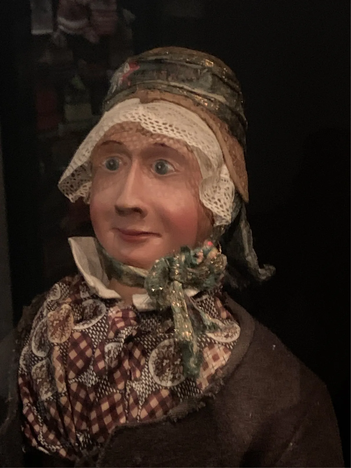 Old wooden doll with painted face, lace bonnet and checked scarf in a dark display