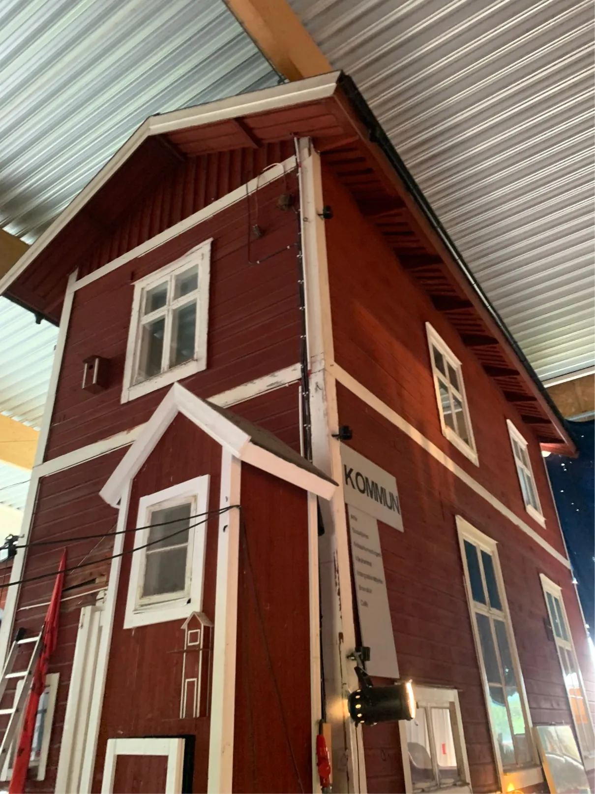 Tall red wooden house with white window frames and a sign reading KOMMUN, seen from below inside a hall
