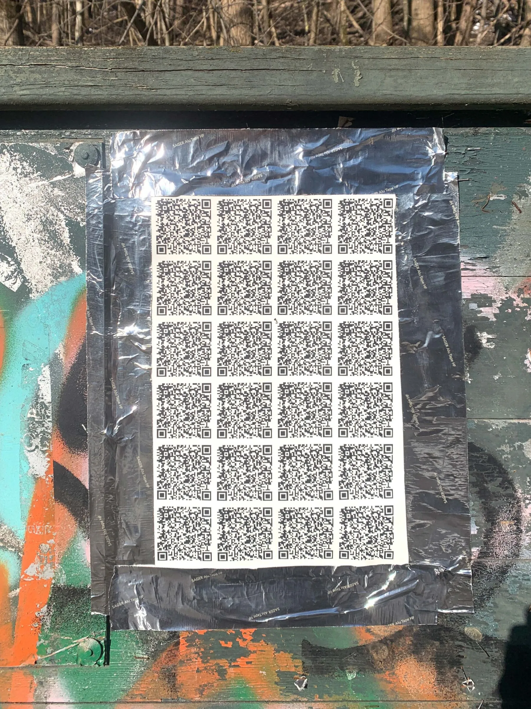 Sheet of identical QR codes taped with foil tape onto a graffiti-covered wooden wall