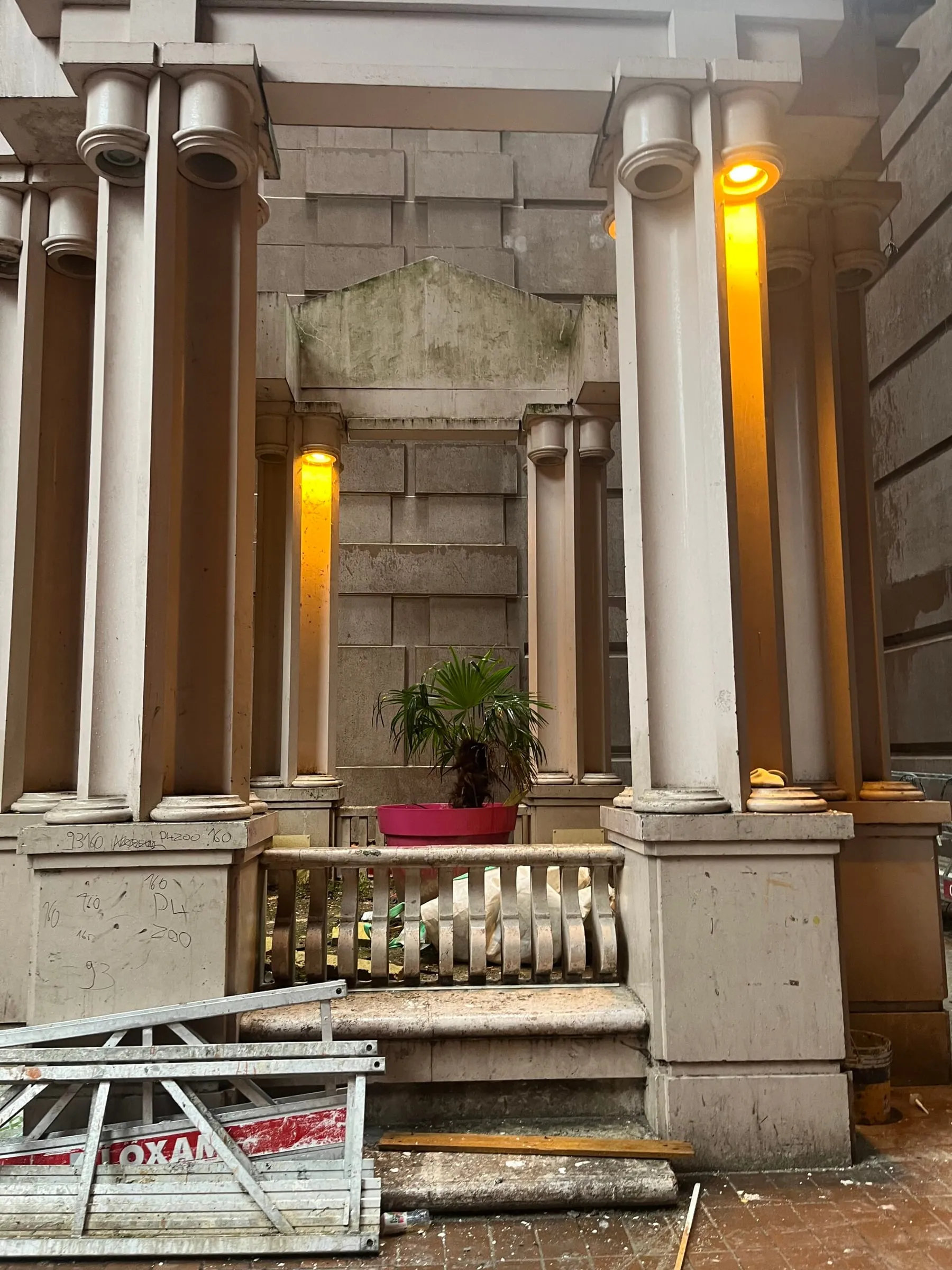 Pale classical columns with warm orange lights, a palm in a pink pot and scaffolding in the foreground