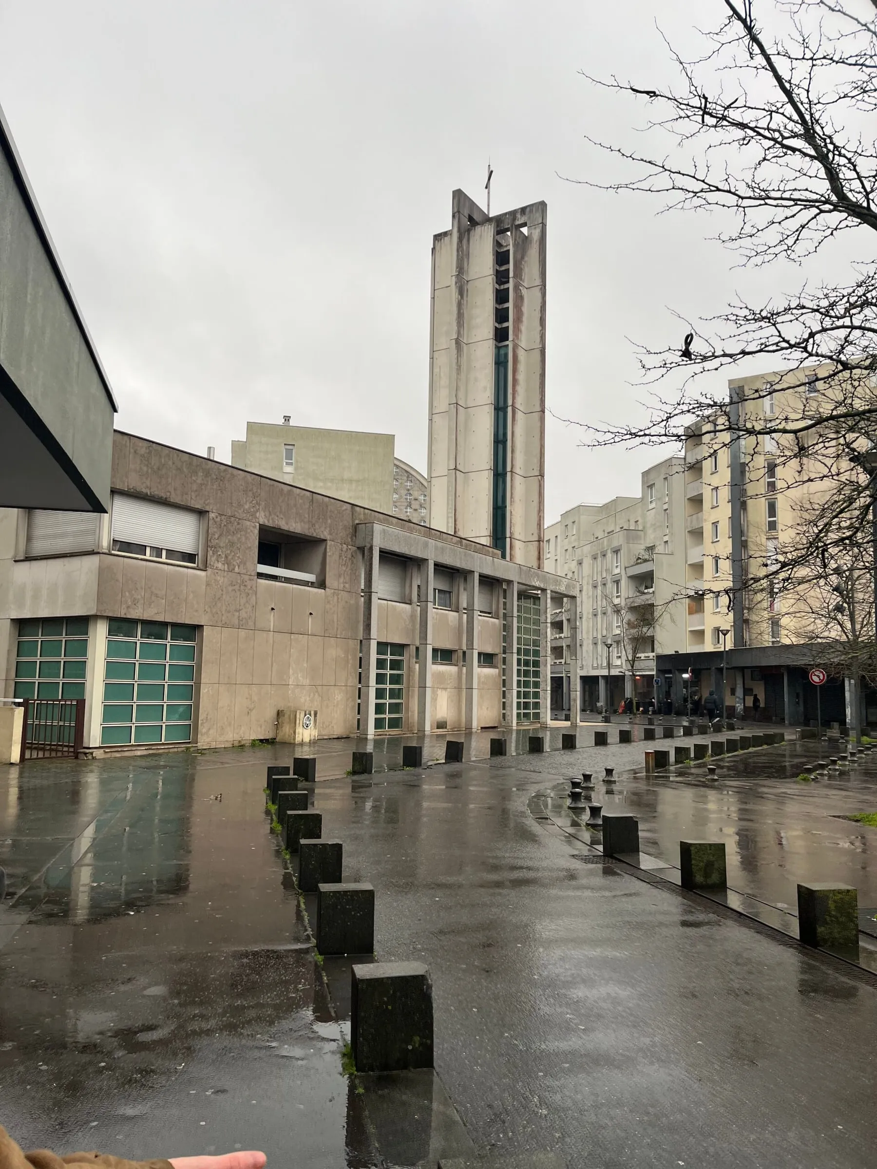 Concrete church tower and apartment blocks on a wet, rainy square lined with stone bollards